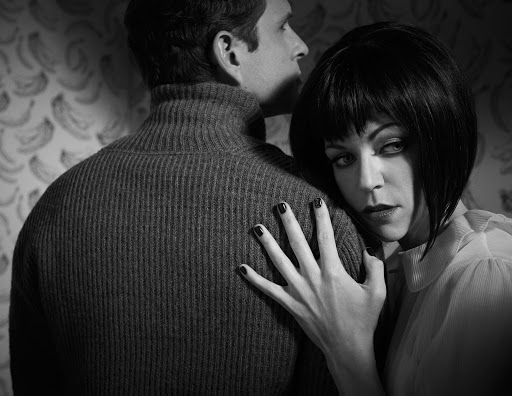 Black and white portrait of a woman with dark bob haircut leaning on a man in a turtleneck sweater against a patterned wall.