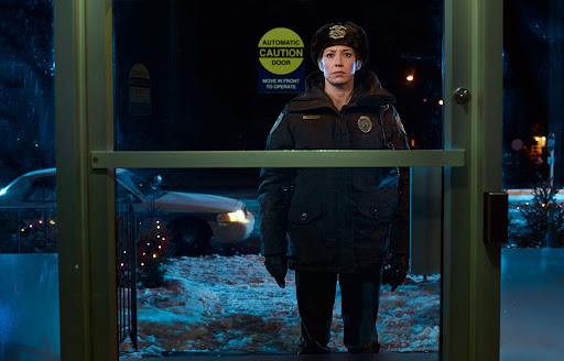 A woman in a police uniform standing at a glass door on a snowy night, with a police car and Christmas tree visible outside.