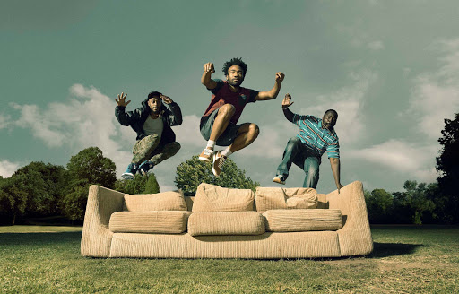 Three men jumping on a sofa outdoors. One man is in mid-air, the other two are on the sofa on green grass with blue sky.