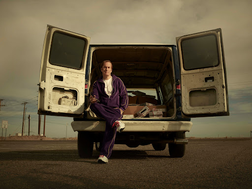 Man in purple tracksuit sits in the open back of a van on an asphalt road, under a cloudy sky, with boxes visible in the van.