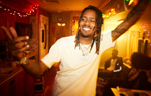 Portrait of a smiling Black man with dreadlocks wearing a white t-shirt and layered necklaces in an indoor setting photographed by Matthias Clamer