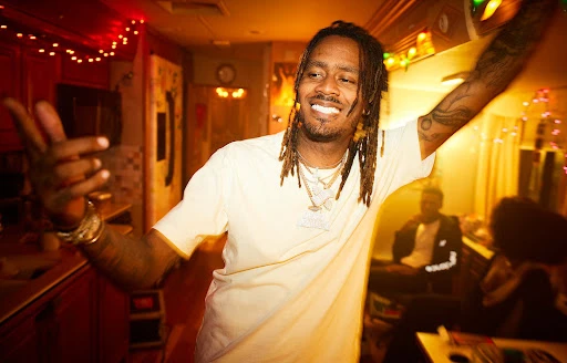 Portrait of a smiling Black man with dreadlocks wearing a white t-shirt and layered necklaces in an indoor setting photographed by Matthias Clamer
