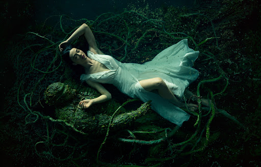 Woman in flowing white dress lies near a mossy sculpture surrounded by vines and dark water, creating a scene of serenity.