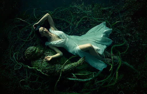 Woman in flowing white dress lies near a mossy sculpture surrounded by vines and dark water, creating a scene of serenity.