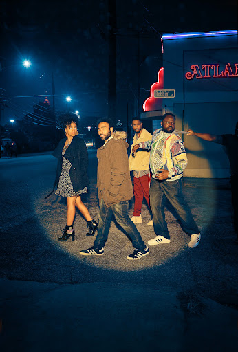 Four people walking on a street at night, illuminated by a circular spotlight. An 'Atlanta' sign glows in the background.