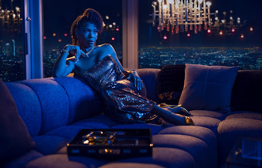 Elegant woman in a gold sequin dress lounges on a sofa in a high-rise apartment with a cityscape view at night.
