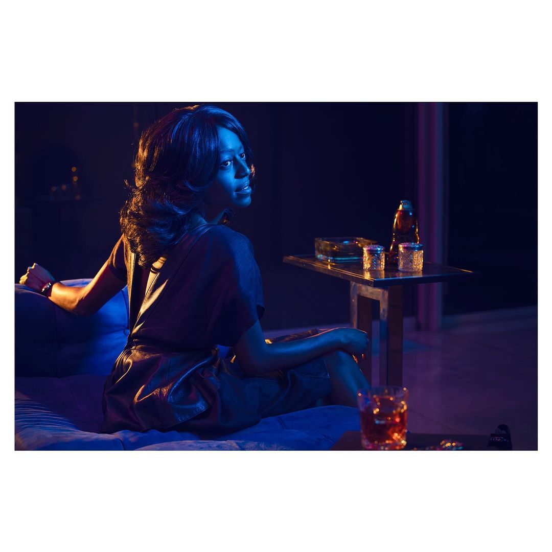Elegant woman lounges on a blue sofa in a dimly lit room with a drink and side table, creating a vintage, intimate, and relaxed ambiance.