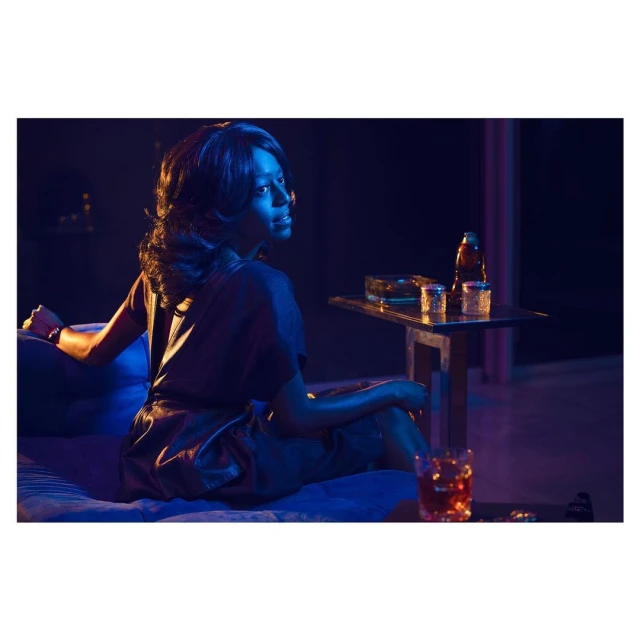 Elegant woman lounges on a blue sofa in a dimly lit room with a drink and side table, creating a vintage, intimate, and relaxed ambiance.