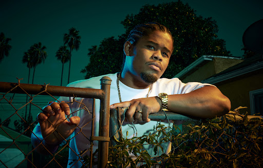 Portrait of an adult male with braids, leaning on a chain-link fence, wearing jewelry and a white t-shirt, with palm trees in the background.
