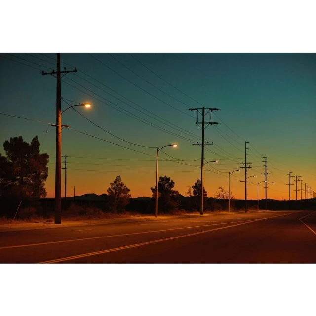 Street at dusk featuring lampposts and telephone poles against a gradient sky of orange, yellow, and blue in an outdoor setting.
