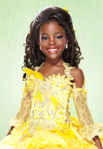 Young African-American girl in a yellow formal gown with lace sleeves and feather accents, posing in front of a soft green background.