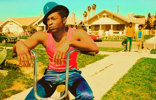 Adult male on bike in residential neighborhood wearing hat and tank top with two background figures on a sunny day.