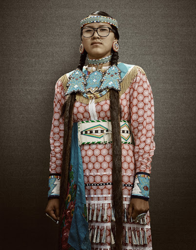 Portrait of an adult woman in Native American traditional clothing with turquoise beadwork and long dark braids in a neutral indoor setting.