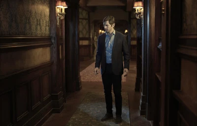 Man in dark blazer and light blue shirt standing in a dimly lit wood-paneled hotel hallway with patterned wallpaper