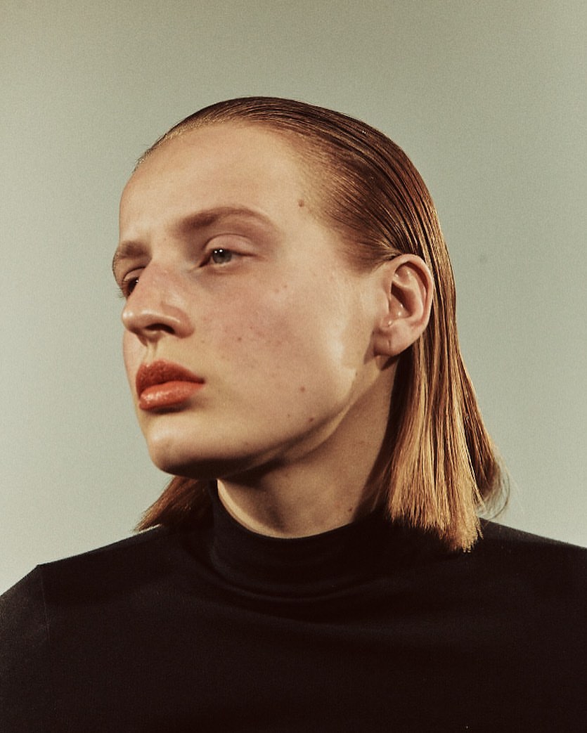 Headshot of a female model with slicked-back red hair and a black turtleneck, set against a neutral studio backdrop.