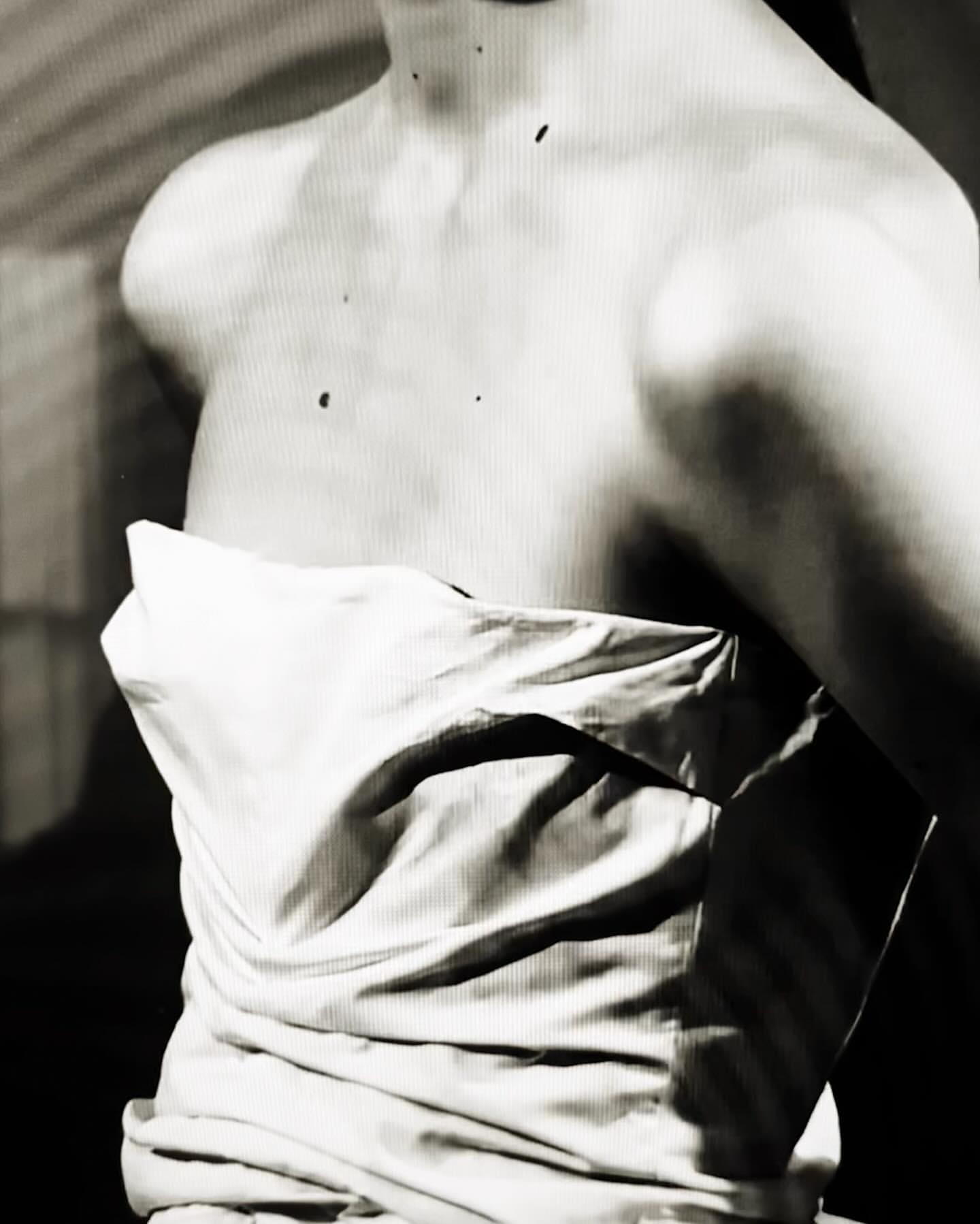 Monochrome portrait of a fair-skinned woman's bare upper body draped with white fabric, set against a muted background.