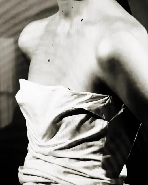 Monochrome portrait of a fair-skinned woman's bare upper body draped with white fabric, set against a muted background.