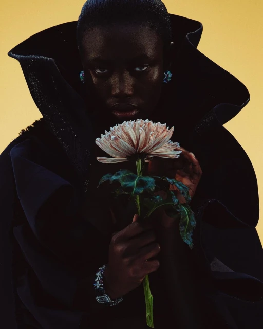 Dark-skinned model in statement collar holds a single large flower against yellow background. Jewelry visible. Beauty and fashion shot.