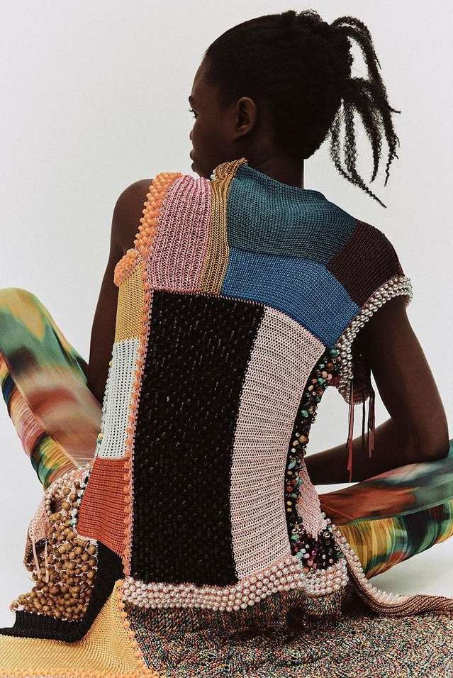 Back view of a Black woman with her hair in a bun, wearing a patchwork knit top with beaded embellishments and multicolor patterned pants.