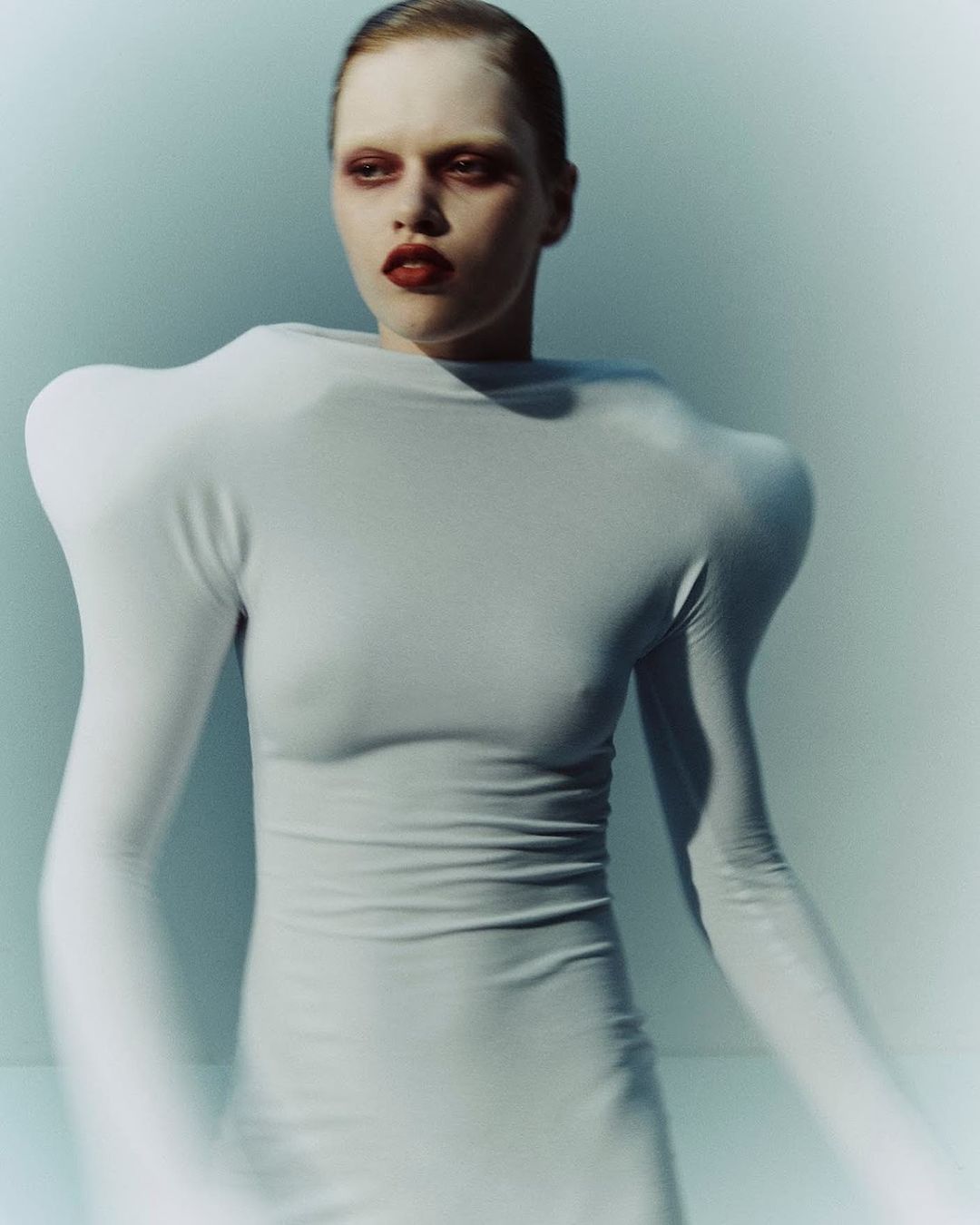 A model with pale skin wears a white dress with shoulder pads in a studio setting. The model has dark eye makeup and lipstick.