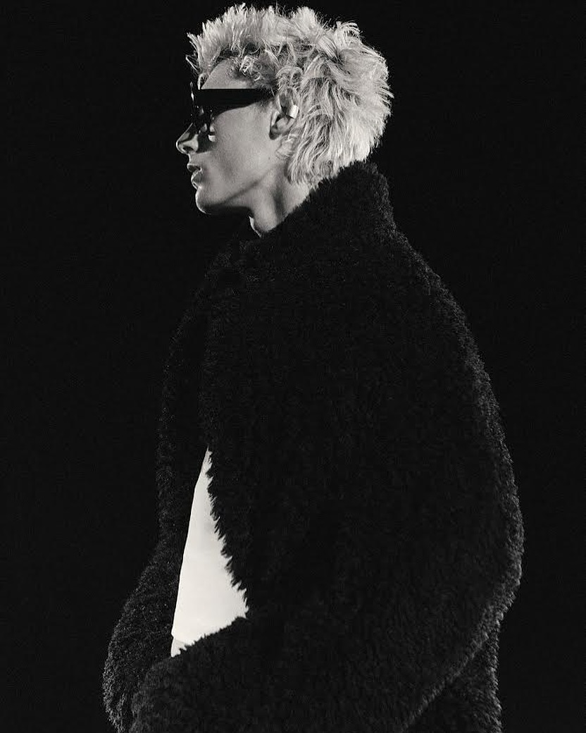 Profile portrait of a person with curly blonde hair wearing dark sunglasses and a thick black textured coat against a dark background.