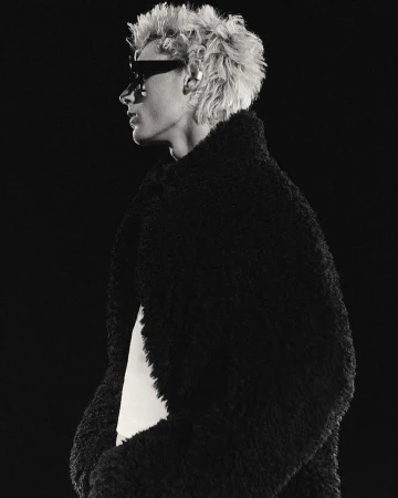 Profile portrait of a person with curly blonde hair wearing dark sunglasses and a thick black textured coat against a dark background.