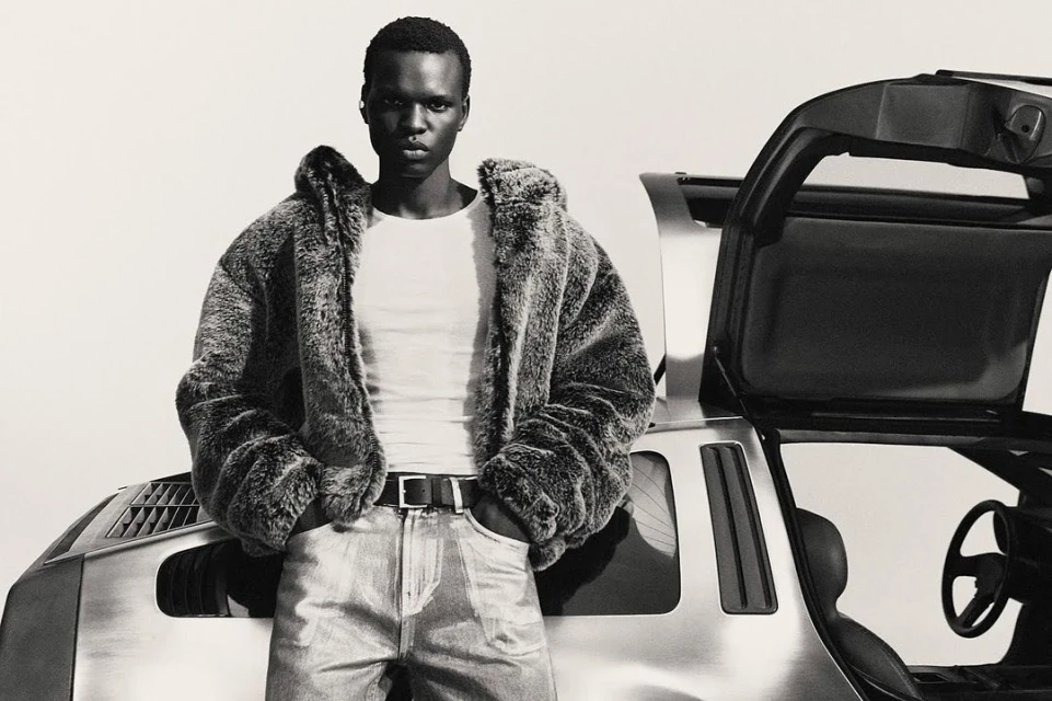 Monochrome portrait of a man in a fur coat standing in front of a Delorean car with gull-wing doors open, creating a retro fashion vibe.