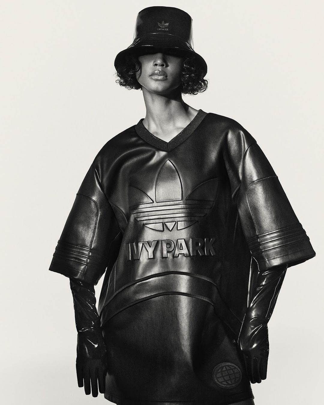 Woman in a shiny black bucket hat, top, and gloves poses in front of a white backdrop. The shirt features the Adidas and Ivy Park logos.