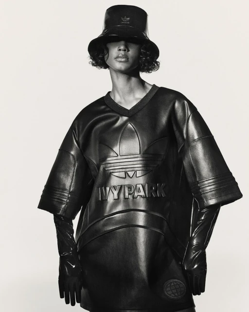 Woman in a shiny black bucket hat, top, and gloves poses in front of a white backdrop. The shirt features the Adidas and Ivy Park logos.