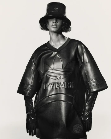 Woman in a shiny black bucket hat, top, and gloves poses in front of a white backdrop. The shirt features the Adidas and Ivy Park logos.