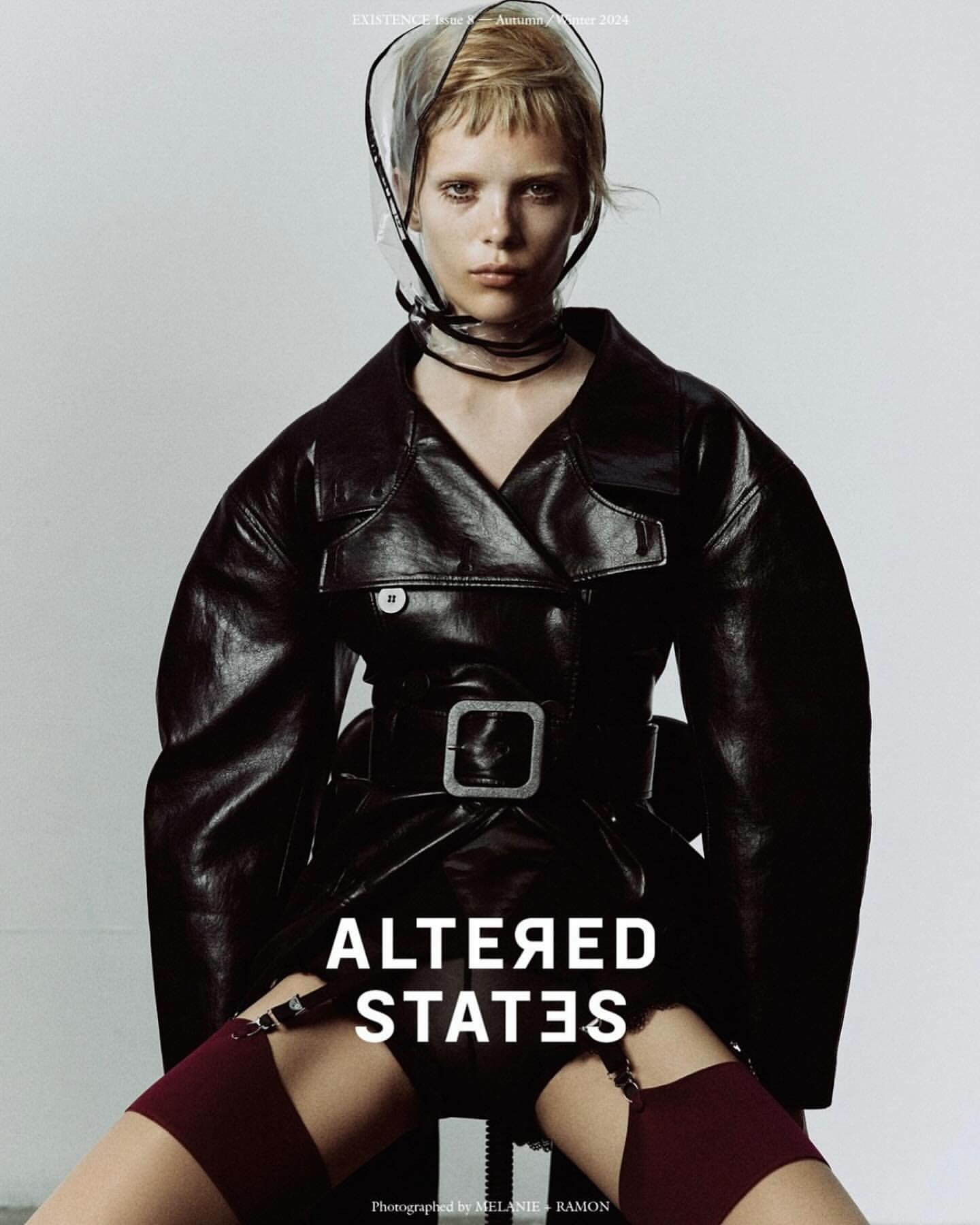 Editorial fashion shot of a woman in a black leather coat, clear plastic hood, stockings, and garter belt against a neutral background.