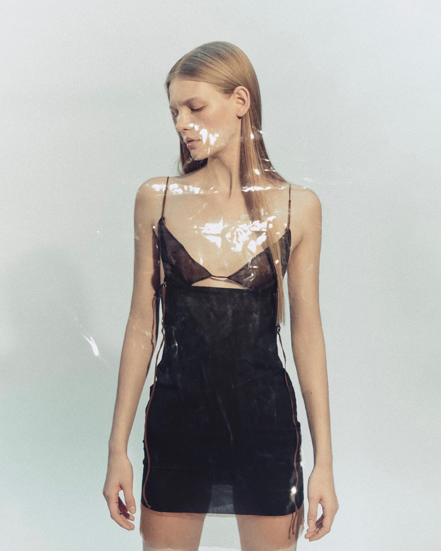 A woman poses in a black slip dress in front of a plain, light background, partially obscured by a clear plastic sheet.