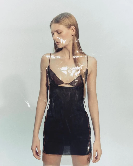 A woman poses in a black slip dress in front of a plain, light background, partially obscured by a clear plastic sheet.