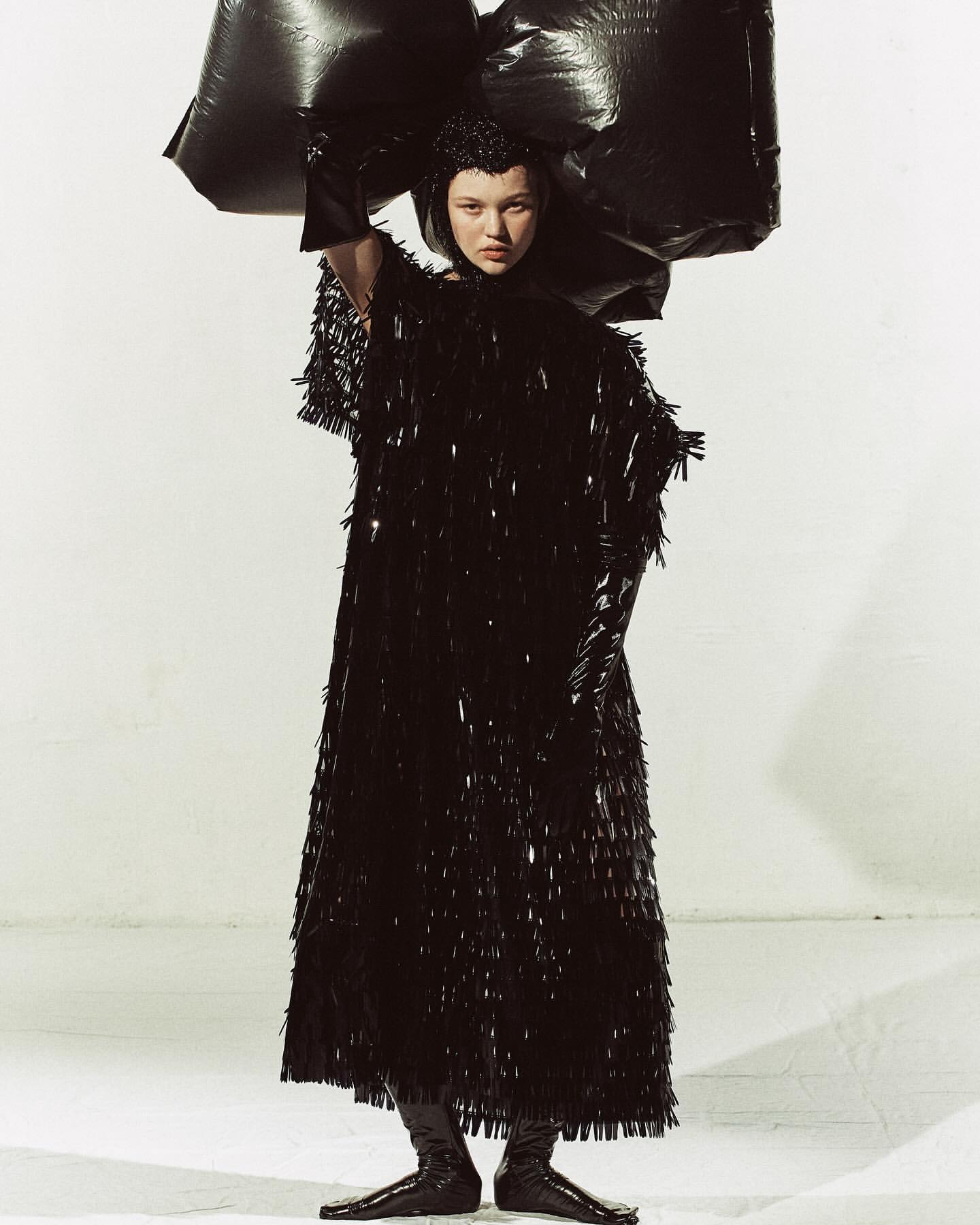 Fashion-forward young woman in a long, shimmery black dress and futuristic headpiece, holding black plastic bags, against a white background.