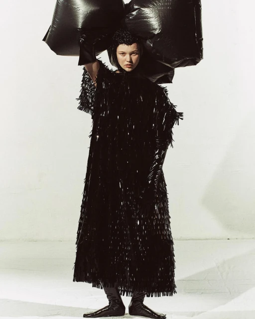 Fashion-forward young woman in a long, shimmery black dress and futuristic headpiece, holding black plastic bags, against a white background.