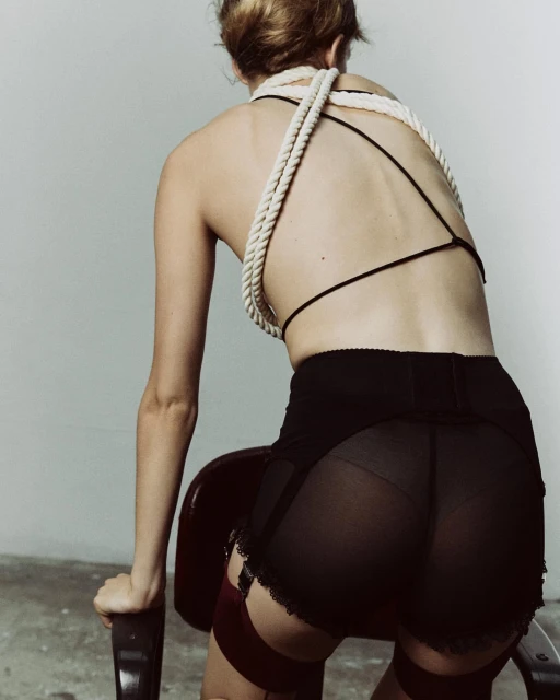 Back view of a woman wearing lingerie with a rope-like top. She is leaning on a chair in a photo studio with a neutral background.