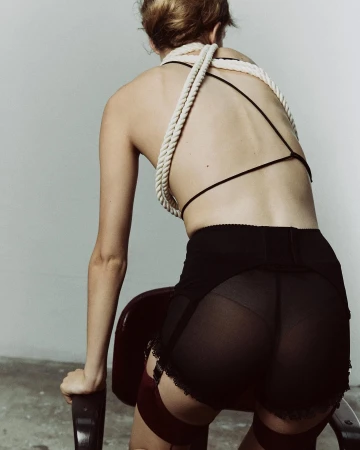 Back view of a woman wearing lingerie with a rope-like top. She is leaning on a chair in a photo studio with a neutral background.