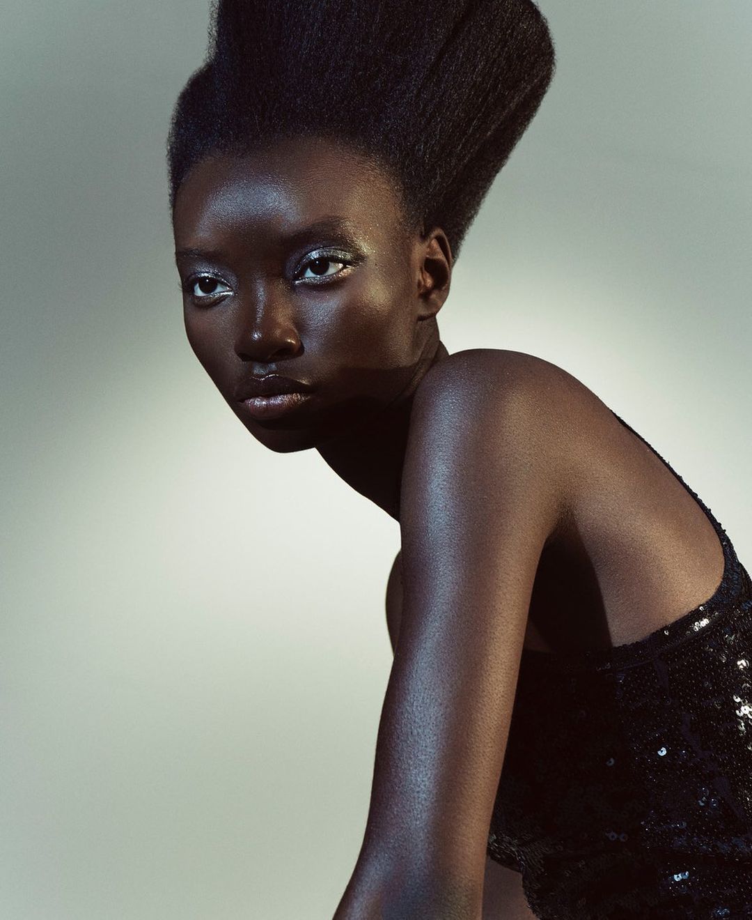 Portrait of a Black woman with dramatic hair posing in a black sequin dress against a grey studio backdrop, gazing off to the side.
