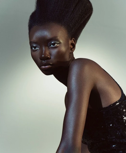 Portrait of a Black woman with dramatic hair posing in a black sequin dress against a grey studio backdrop, gazing off to the side.