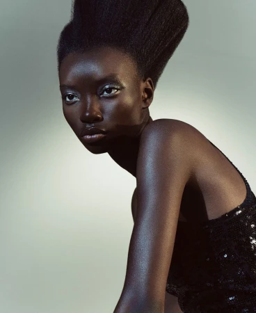Portrait of a Black woman with dramatic hair posing in a black sequin dress against a grey studio backdrop, gazing off to the side.