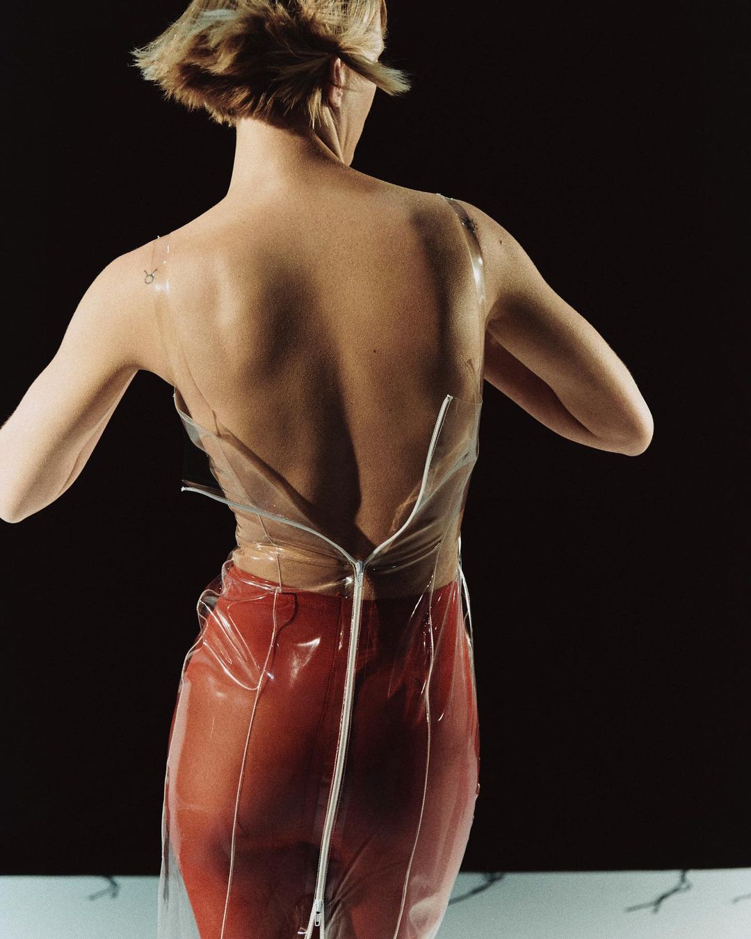 Back view of a slender adult female wearing a transparent top and red skirt against a stark black background in a studio setting.