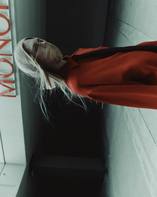 Woman in orange shirt, black shoulder strap, and blonde hair in a shadowy indoor setting with MONO text on a sign.