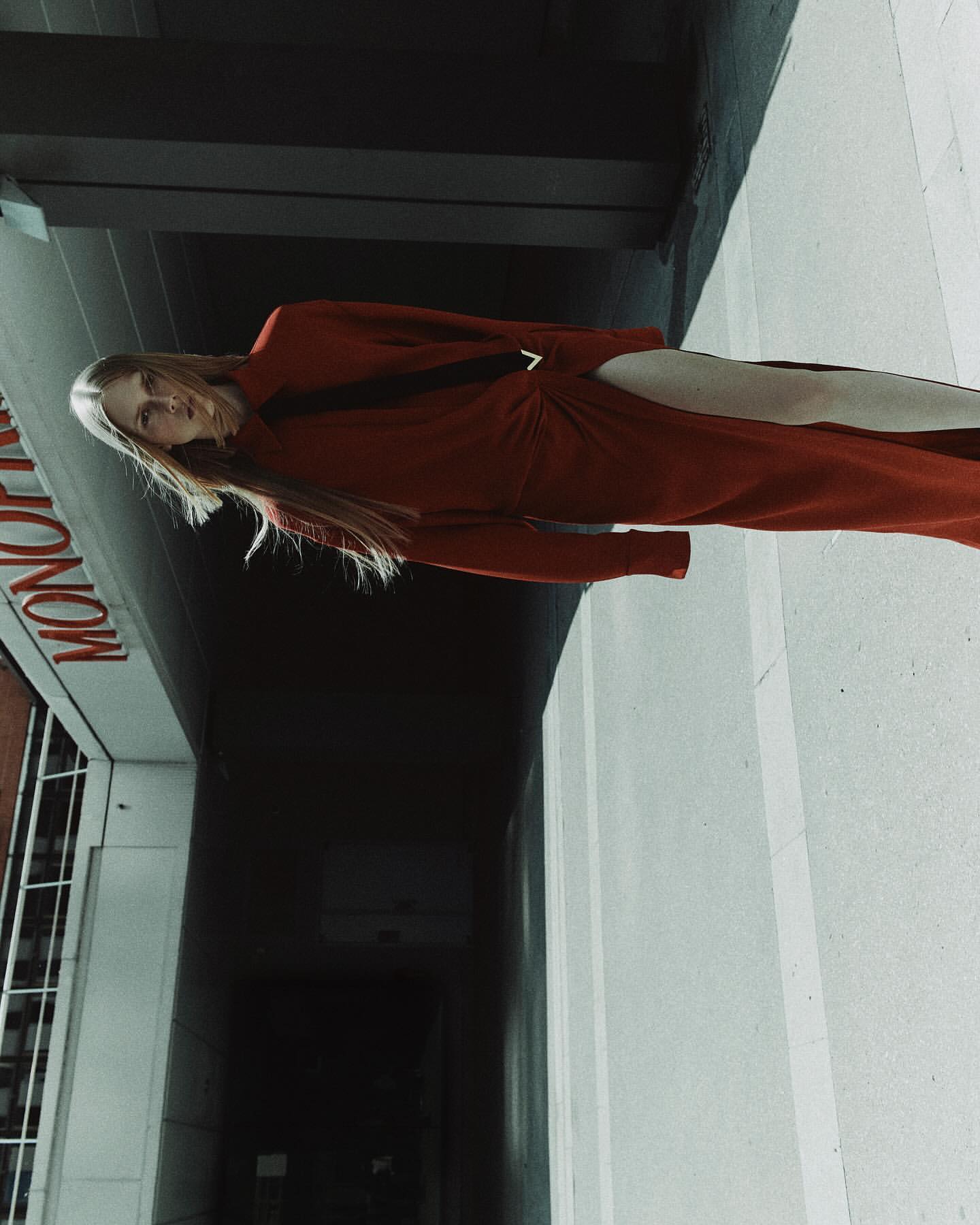 A blonde female model wearing a long red dress standing outdoors under a concrete structure with a sign reading MONO visible.