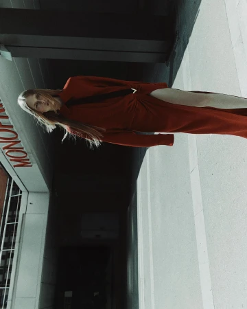A blonde female model wearing a long red dress standing outdoors under a concrete structure with a sign reading MONO visible.
