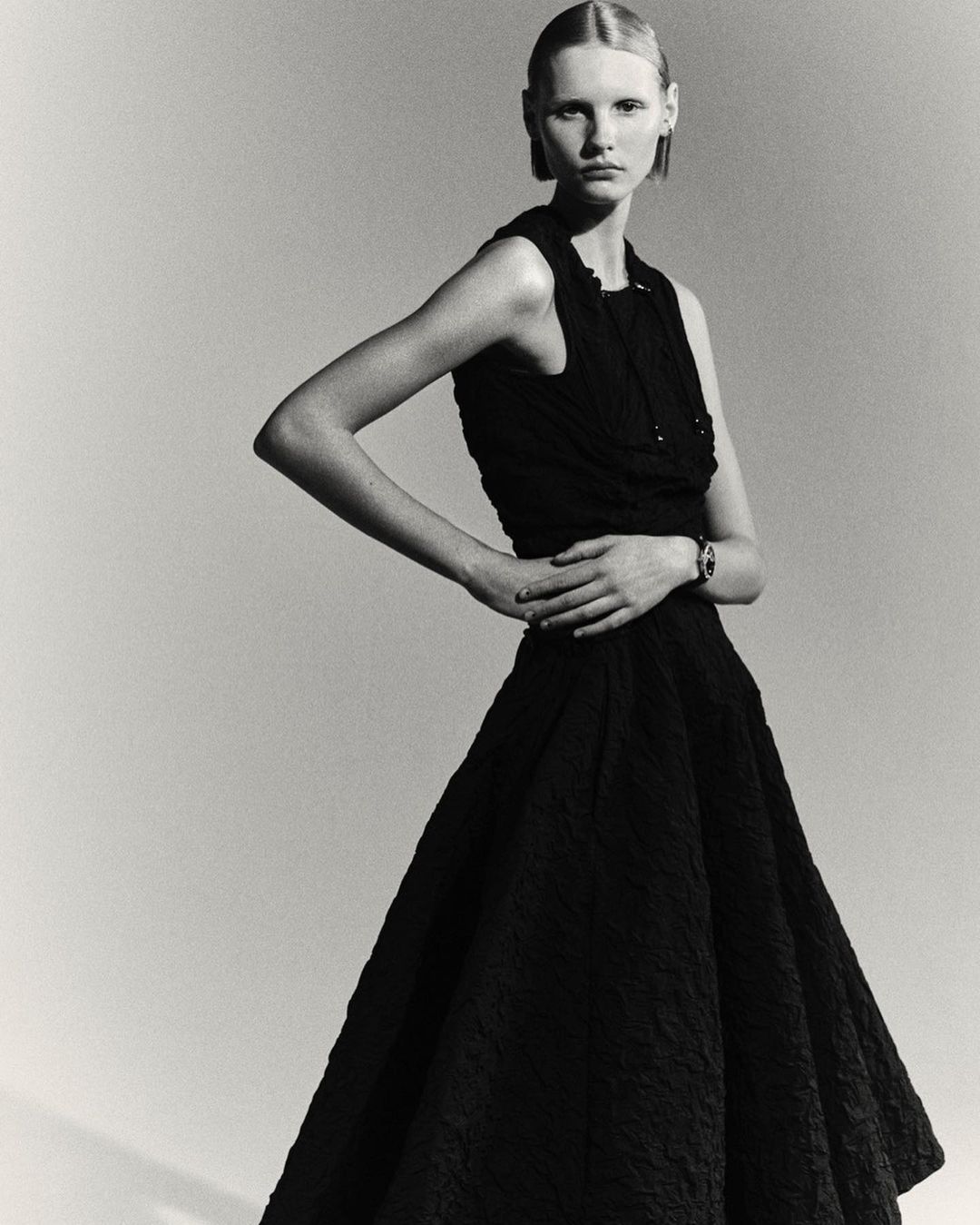 Model in a black sleeveless dress posing against a plain grey studio backdrop with hands on hips