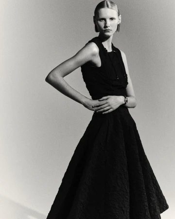 Model in a black sleeveless dress posing against a plain grey studio backdrop with hands on hips