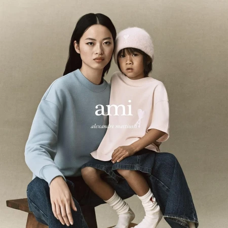 A fashionable campaign image features a woman and child modelling AMI apparel. They wear casual clothing, set against a neutral backdrop.
