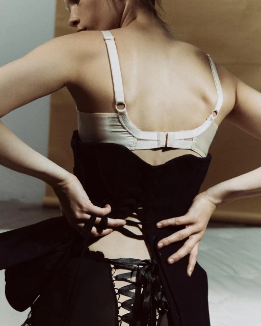 A young adult female wears a black corset with satin ribbons and ties it behind her back, shot in front of a beige-toned backdrop.