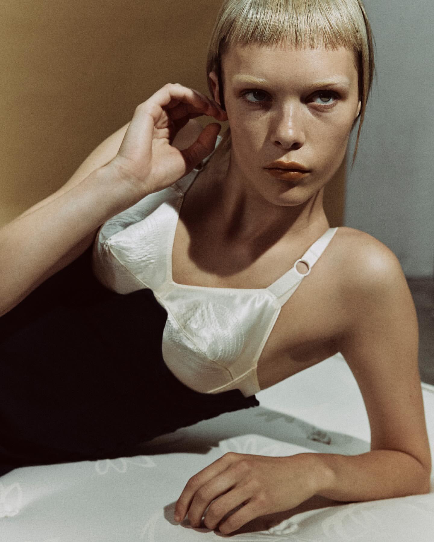 A fair-skinned woman with short blonde hair, wearing a white bra and black top, lies on a white surface against a tan and gray backdrop.