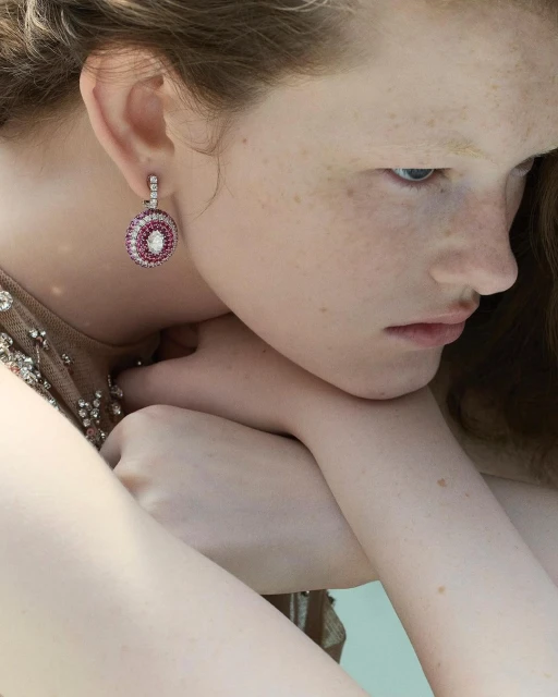 Close-up of a fair-skinned woman wearing round, ornate drop earrings with pink and white stones, showcasing jewelry.